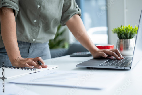 Businesswoman sitting at desk on couch in workplace or at home working on laptop and analyzing data on charts and graphs and writing on papers to make business plan and strategies for company
