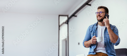 Handsome bearded man leaning on office desk, drinking coffee and talking on the phone.