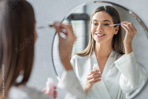 A happy millennial woman in a silk robe applies skin serum to her face with a dropper while looking at her reflection in the bathroom. This marks her beauty routine at home.