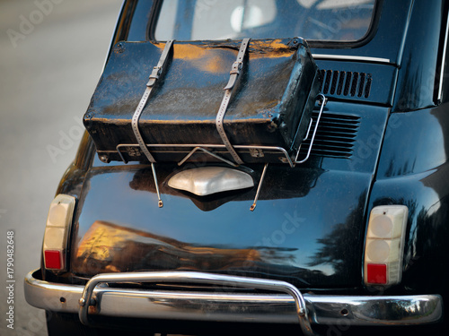Rear view of a black vintage car with a luggage rack and an old suitcase strapped on it.