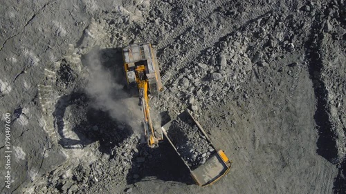 Aerial top-down view of an excavator loading rocks into a dump truck at an open-pit mine. Industrial drone footage showing heavy machinery working in a dusty environment, highlighting extraction