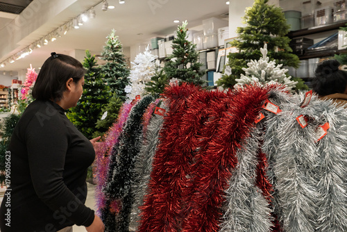 A woman browses colorful tinsel garlands displayed in a store aisle decorated with artificial Christmas trees. The festive decorations suggest holiday shopping season.