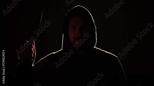 Mysterious man in a dark hoodie holding a large kitchen knife, illuminated by dramatic low light. Suspenseful and cinematic scene symbolizing danger, crime, or thriller concept.