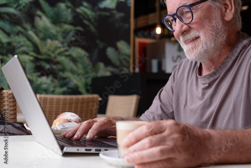 Tableau sur toile Smiling senior man sits in a cafe having break with coffee and croissant while using his laptop