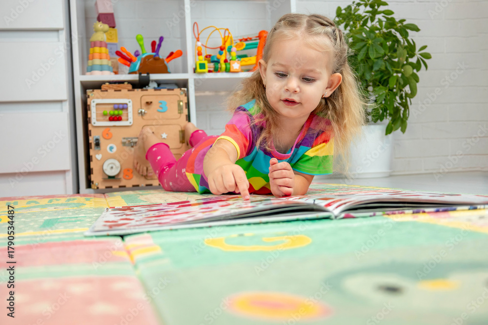Fototapeta premium Toddler girl in bright clothes playing and learning with a book on a colorful floor mat at home.