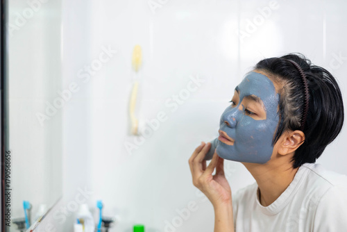 Asian woman applies clay facial mask in front of bathroom mirror. Skincare routine promotes clean skin and natural beauty. A refreshing beauty treatment for detoxifying and rejuvenating the face.