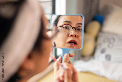 Beautiful Asian woman applies lipstick in front of handheld mirror. Close-up reflection shows candid beauty routine of self care, natural skin, and confident expression during personal makeup moment.