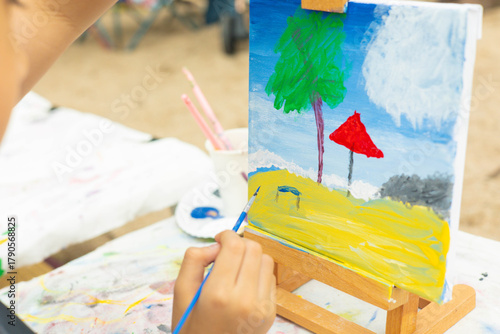 Close-up shot of a child's hand applying paint to a canvas. The colorful artwork depicts a beach scene with a tree and umbrella, showing childhood creativity, art education, and imaginative hobbies.