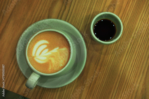 A top-down view of a latte with foam art in a ceramic cup and saucer, placed next to a small cup of dark liquid, both resting on a wooden table with a visible grain.