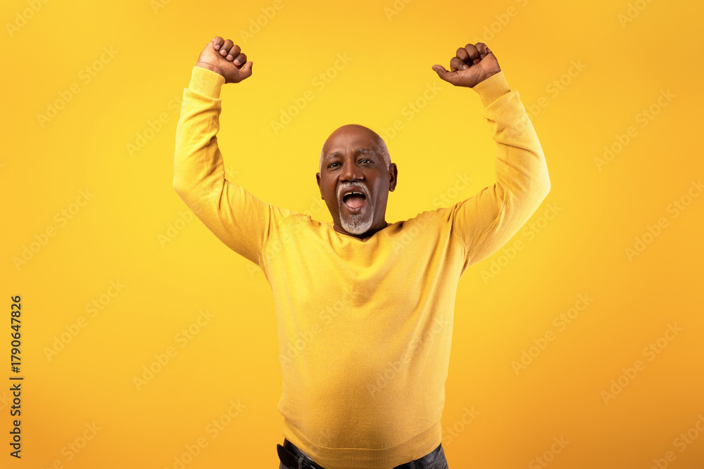 Fototapeta premium An overjoyed senior black man expresses excitement by shouting and raising his arms in triumph. He is set against a bright orange background, showcasing his happiness and enthusiasm for success.