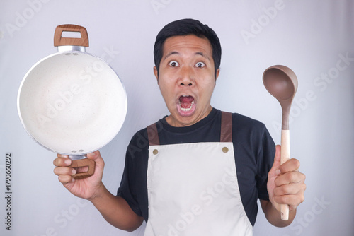 Funny excited expression asian man chef wearing apron holding pan and soup spoon on white background
