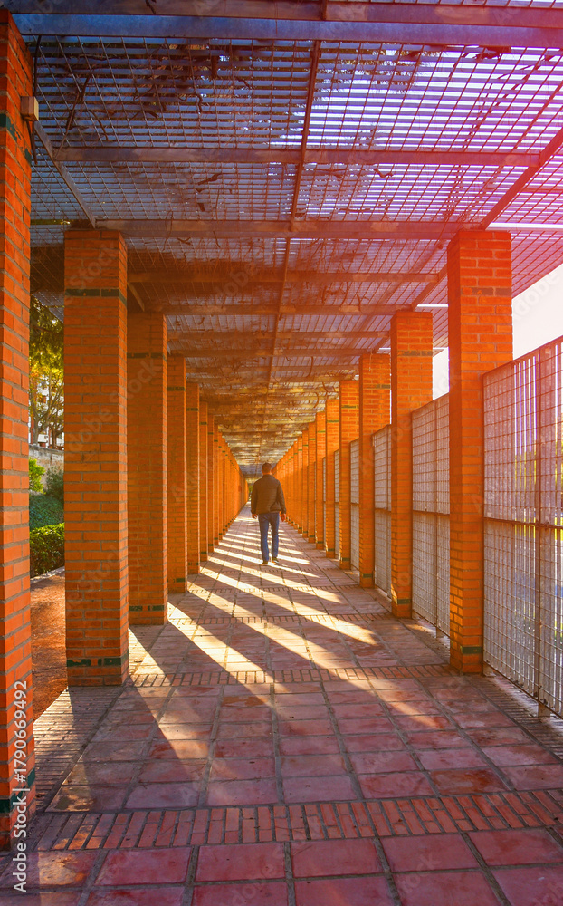 Fototapeta premium Man in Brick Corridor With Light Streaming. Walkway with warm sunlight illuminating brick pillars and shadows in a peaceful park setting. Person silhouette walks in long corridor with brick pillars.