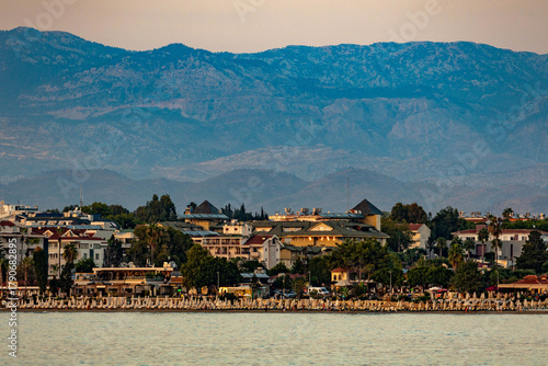 Fototapeta Naklejka Na Ścianę i Meble -  Sunset view of Side's coastline with white umbrellas, promenade cafes and hotels against forested mountains in golden evening light. Antalya, Turkey.


