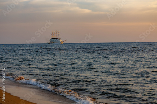 Fototapeta Naklejka Na Ścianę i Meble -  Sunset over Mediterranean Sea with gentle waves on sandy beach and a pirate-themed tourist ship sailing on horizon, illuminated by warm evening light. Antalya, Turkey.

