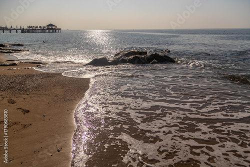 Fototapeta Naklejka Na Ścianę i Meble -  Morning beach scene with backlight creating sparkle on wet sand and wave foam, rocky outcrops and overwater sundeck against sun path on horizon. Sorgun, Antalya, Turkey.

