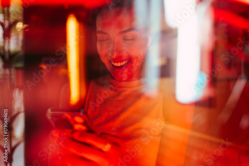 Smiling woman lit by warm neon reflections uses smartphone, capturing joy and connection in a tech-immersed moment, symbolizing the emotional side of digital interaction.