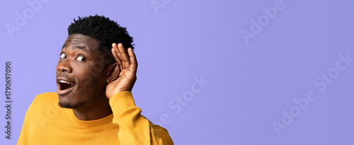 A young man wearing a yellow sweater is looking surprised while holding his hand to his ear. He appears eager to hear something interesting or exciting against a solid purple background.