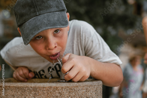 Konstfotografi Boy in a gray cap and white t-shirt drinking water from a public fountain on a sunny summer day