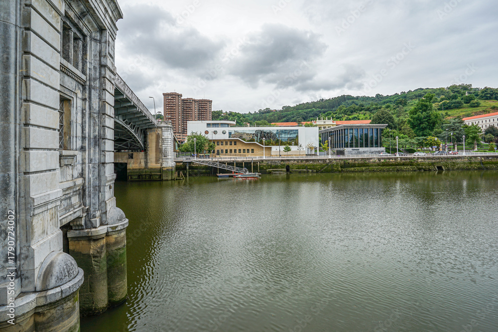 Fototapeta premium View of the embankment from the river in the city of Bilbao, Spain. A large stone bridge is visible on the left side The photo was taken in cloudy weather, with the sky covered in clouds