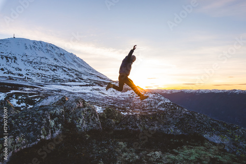 Traveler jumps from rocks at sunset near Gaustatoppen Mountain in Norway. Golden light, snow peaks, and open sky capture pure joy and freedom of adventure