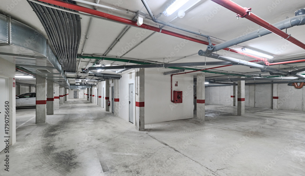 Obraz premium Clean and spacious underground parking garage with ventilation ducts, pipes, and red safety markings on concrete walls and columns