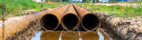 Rusty distribution pipe trio in dried canal with shallow reflective water, sunlit industrial drainage scene conveying decay and quiet atmosphere