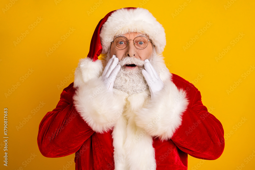 Naklejka premium Santa Claus in red suit with white beard and glasses surprised holding cheeks against bright yellow background festive holiday scene