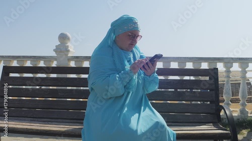 A young Muslim woman in a blue hijab sits on a bench by the sea on the embankment