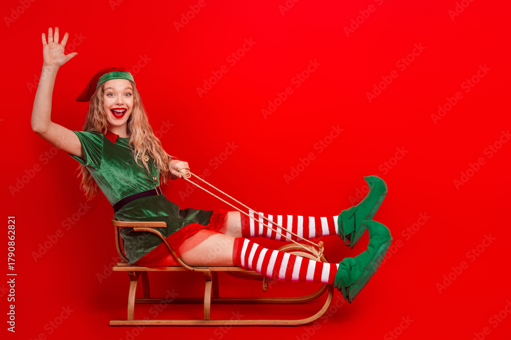 Obraz premium Joyful elf celebrating Christmas season on a sled, wearing festive costume against red background in a cheerful pose