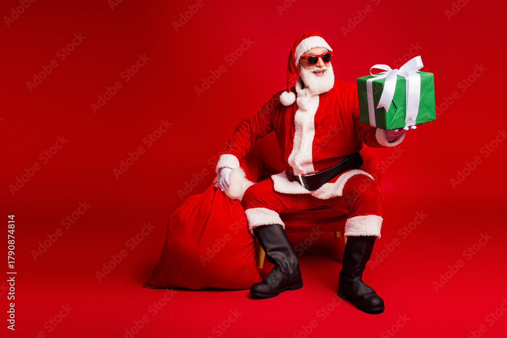 Obraz premium Santa Claus in red suit offering green gift against bold red background for christmas season celebration and holiday sales