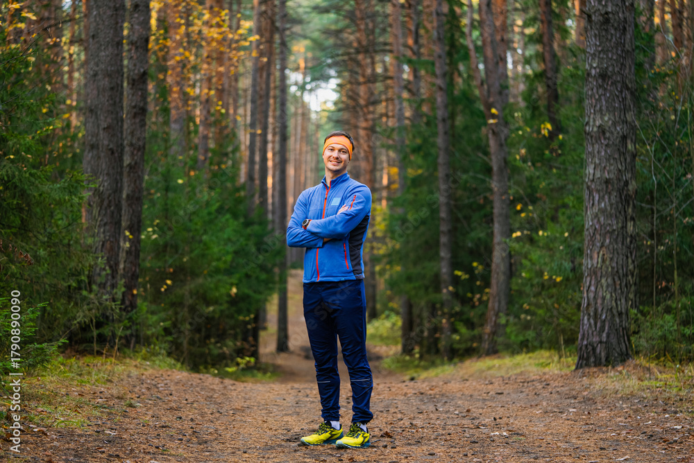 Obraz premium Man standing on a dirt trail in a forest, smiling, dressed in sportswear with arms crossed, enjoying an active outdoor lifestyle and healthy fitness routine during autumn season