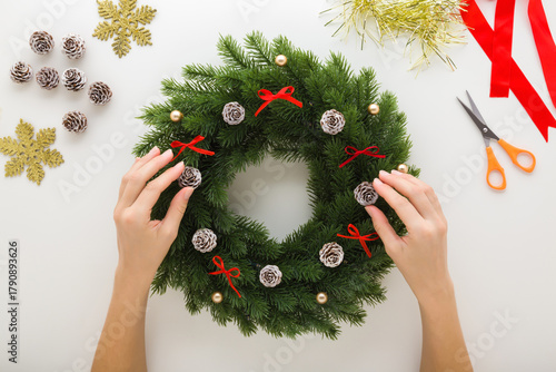 Young adult woman hands making green fir wreath with decoration on light gray table background. Creating Christmas door wreath. Flat lay. Closeup. Point of view shot. Top down view.