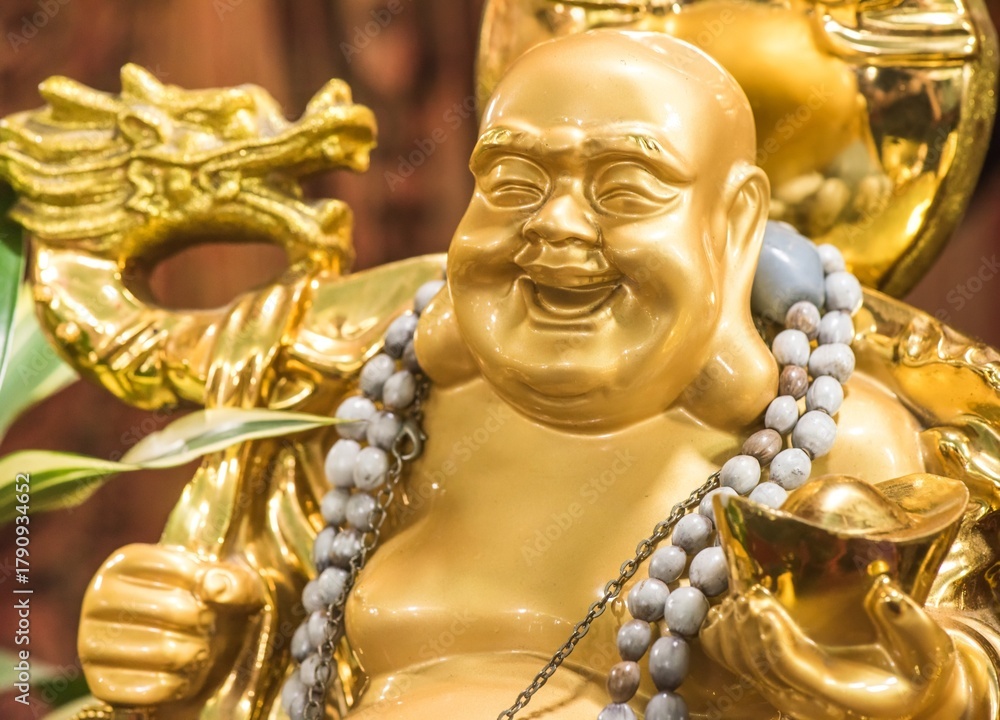 Fototapeta premium Close up of a golden Laughing Buddha statue (Budai or Hotei) holding a gold ingot. Figure symbolizes wealth, happiness, and good fortune in Feng Shui and Buddhism.