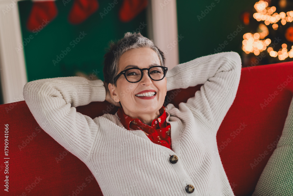 Fototapeta premium Smiling senior woman in cozy winter cardigan relaxing at home near festive Christmas decorations and a lit-up tree