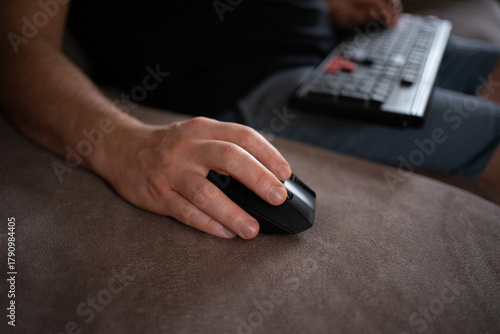 Close-up of a person using a wireless computer mouse on a sofa while holding a keyboard. Cozy remote work, gaming, or home office concept with modern technology and relaxed atmosphere