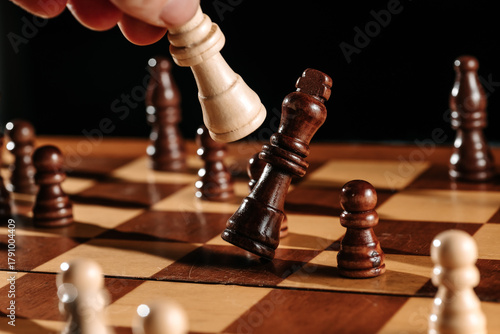 A hand delivers the final capture as a white piece knocks over the dark king—decisive endgame moment frozen on the wooden chessboard.