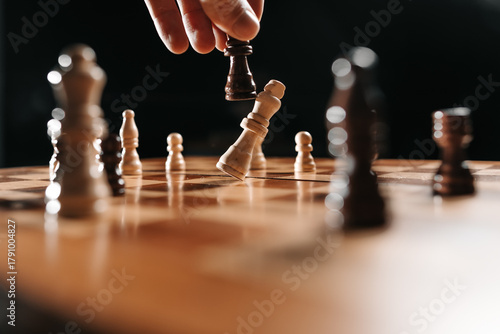 Human hand holds a black chess piece as the white king falls, illustrating strategy, decisive move, leadership and competitive mindset.