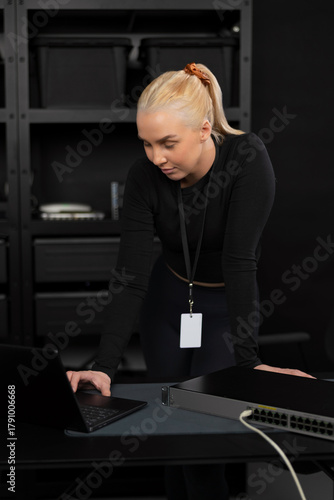 Female IT network technician in her twenties reviewing laptop beside network switch in server room