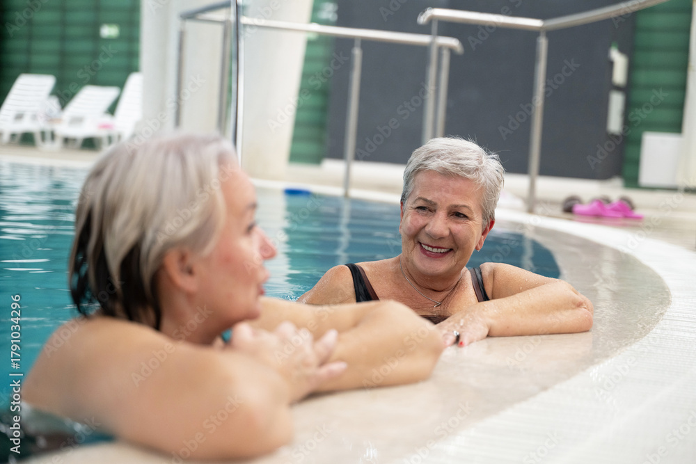 Naklejka premium Two smiling senior women enjoying leisure time in an indoor swimming pool, leaning on the poolside. Concept of friendship, wellness, active lifestyle, relaxation, and healthy aging.