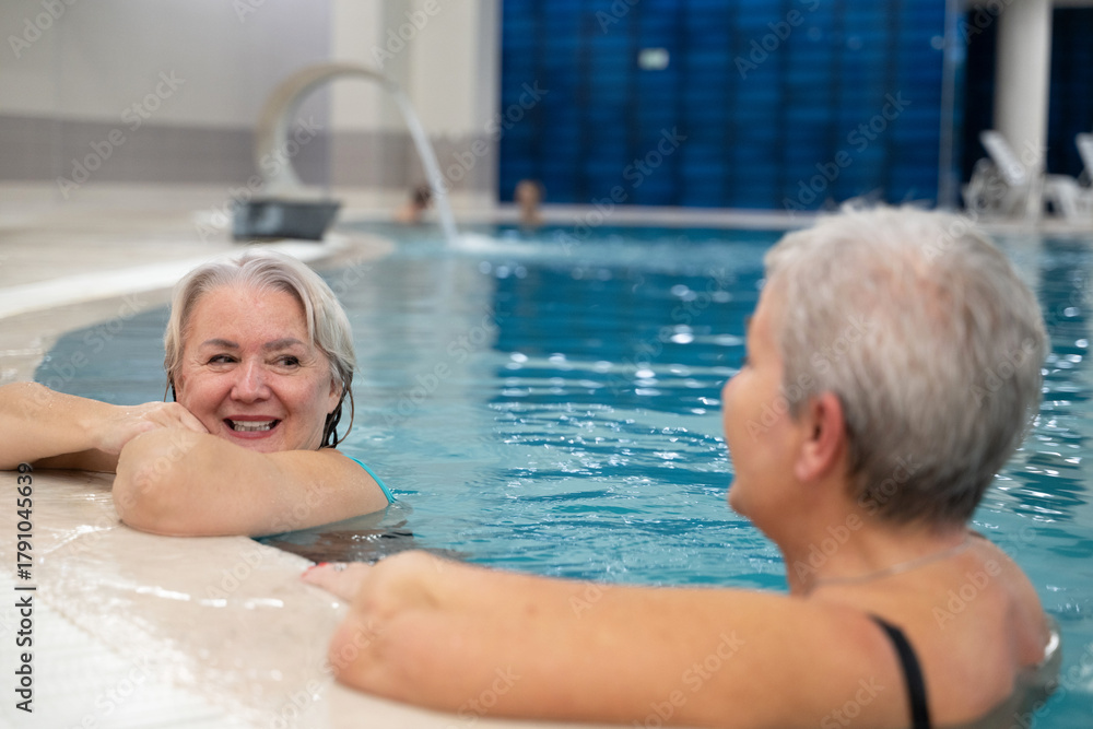 Fototapeta premium Two smiling senior women enjoying leisure time in an indoor swimming pool, leaning on the poolside. Concept of friendship, wellness, active lifestyle, relaxation, and healthy aging.