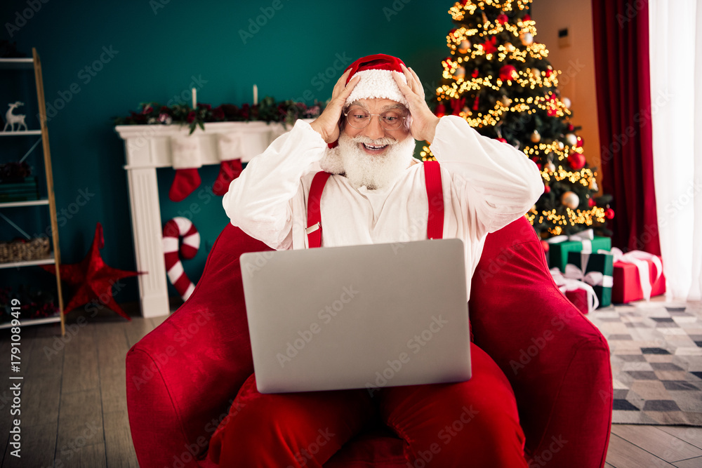 Naklejka premium Santa at home uses a laptop in a cozy living room with a Christmas tree gifts and festive lights creating a joyful holiday scene