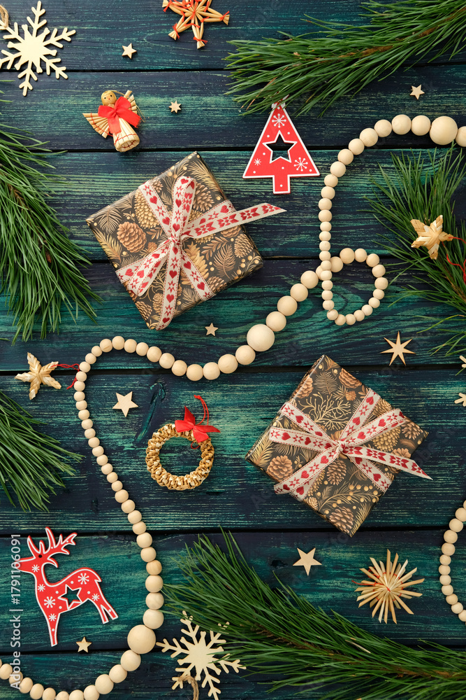 Fototapeta premium Christmas gifts with pinecone wrapping, red ribbon, pine branches, reindeer, angel, wreath, and festive ornaments on dark teal wooden surface.