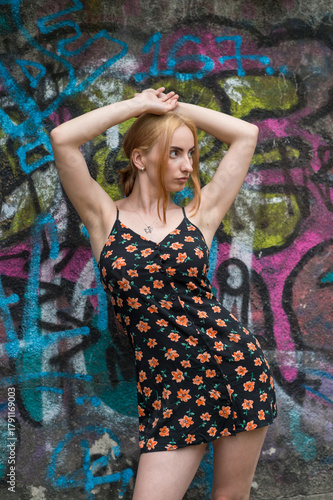 Blonde sensual woman with long legs posing confidently in front of a colorful graffiti wall, wearing a short floral dress. Urban fashion meets vibrant street art in this expressive portrait.