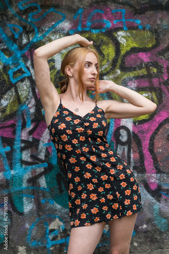 Blonde sensual woman with long legs posing confidently in front of a colorful graffiti wall, wearing a short floral dress. Urban fashion meets vibrant street art in this expressive portrait.
