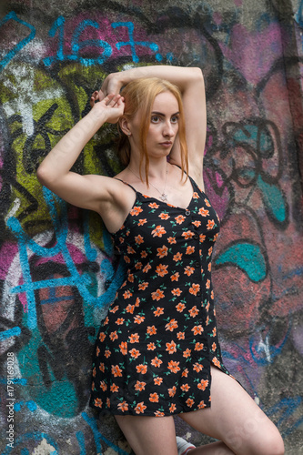 Blonde sensual woman with long legs posing confidently in front of a colorful graffiti wall, wearing a short floral dress. Urban fashion meets vibrant street art in this expressive portrait.