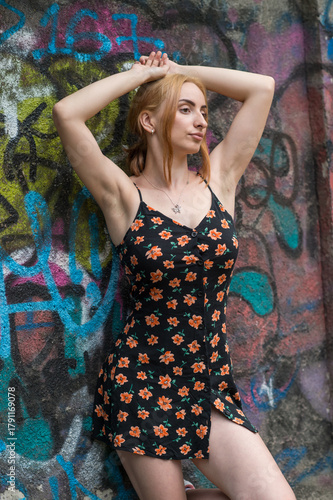 Blonde sensual woman with long legs posing confidently in front of a colorful graffiti wall, wearing a short floral dress. Urban fashion meets vibrant street art in this expressive portrait.