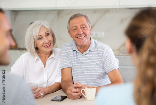Canvas Print Smiling elderly parents and adult children sit at the table with tea and talk ab