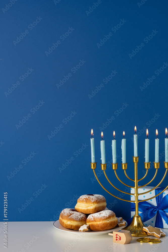 Fototapeta premium A vertical festive Hanukkah scene featuring a gold menorah with lit candles, a plate of powdered sufganiyah donuts, and wrapped gifts against a rich blue backdrop conveying