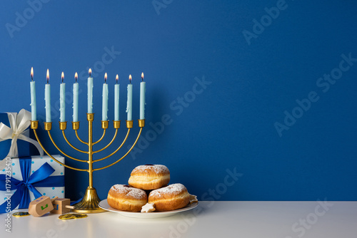 Vibrant Hanukkah setting featuring a gold menorah with lit candles, wrapped gifts, a plate of sufganiyot, and gelt on a white surface against a deep blue wall