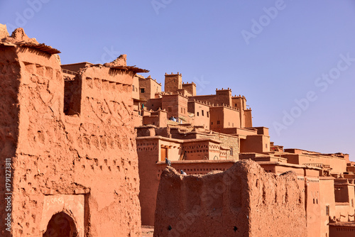 This is a view of the ancient Ksar of Ait ben Haddou. The traditional mud brick buildings are stacked on a hillside with a clear blue sky above, showing the texture of the architecture